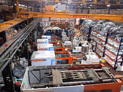 A row of horizontal injection molding presses on the production floor, with material stored on racking alongside and an overhead travelling crane above